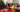 Young adult women in red pants and a yellow sweater relaxing on the floor in front of a Christmas tree.