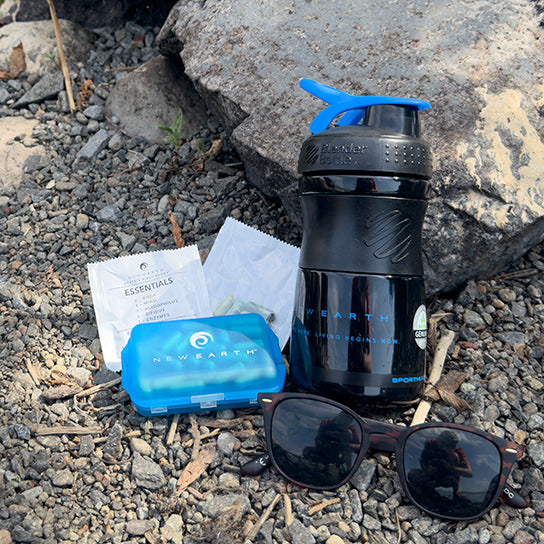 A New Earth Mixer Botttle sitting on the ground next to a two packets of Essentials, a New Earth pill pack, and a pair of sunglasses