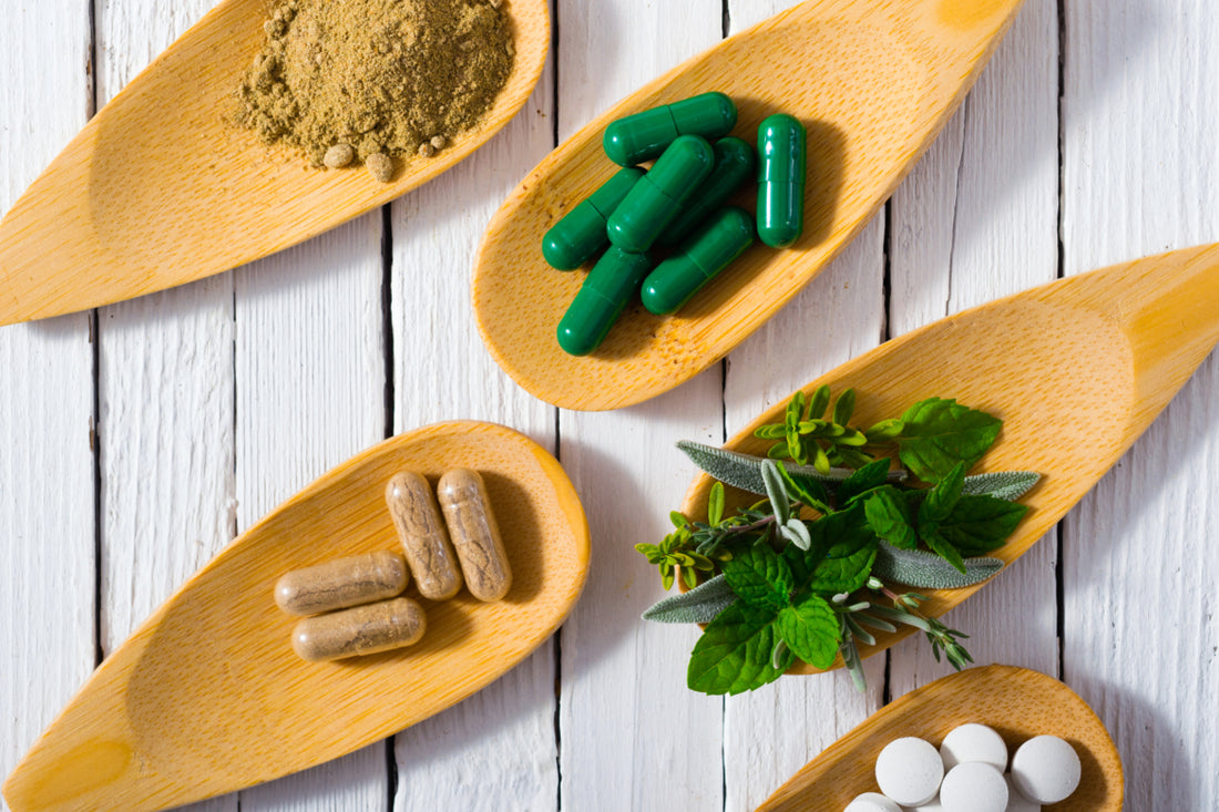 Five wooden spoons laying on a table. Each spoon is holding a different item including: brown food powder, green supplement capsules, brown supplement capsules, green herbs, and white pills.
