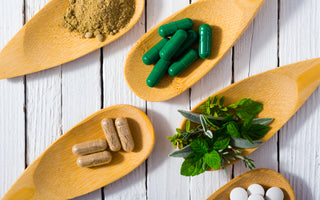 Five wooden spoons laying on a table. Each spoon is holding a different item including: brown food powder, green supplement capsules, brown supplement capsules, green herbs, and white pills.