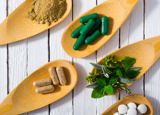 Five wooden spoons laying on a table. Each spoon is holding a different item including: brown food powder, green supplement capsules, brown supplement capsules, green herbs, and white pills.