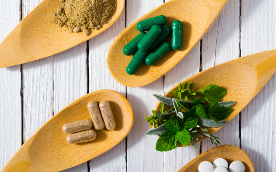 Five wooden spoons laying on a table. Each spoon is holding a different item including: brown food powder, green supplement capsules, brown supplement capsules, green herbs, and white pills.