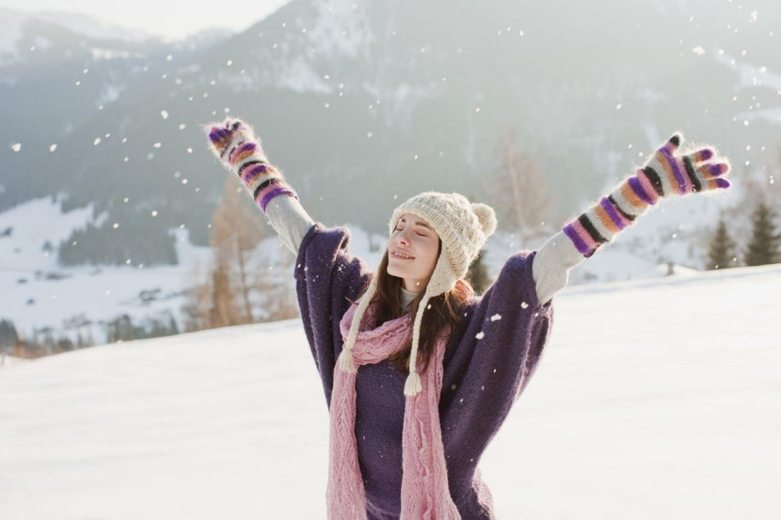 Young woman dressed in winter gear standing in the snow with her arms spread out above her head.