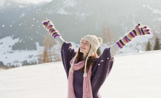 Young woman dressed in winter gear standing in the snow with her arms spread out above her head.