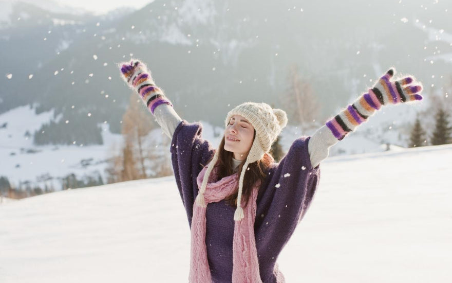 Young woman dressed in winter gear standing in the snow with her arms spread out above her head.