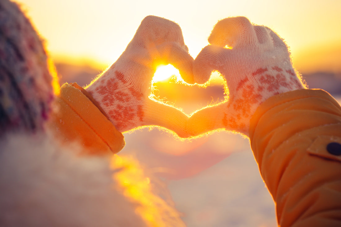 Hands wearing winter gloves are held up with fingers positioned to form a heart in front of the sunset.