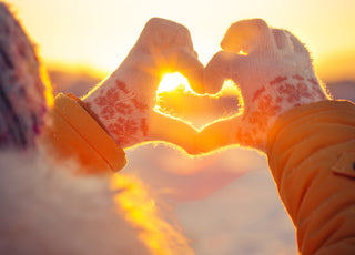 Hands wearing winter gloves are held up with fingers positioned to form a heart in front of the sunset.