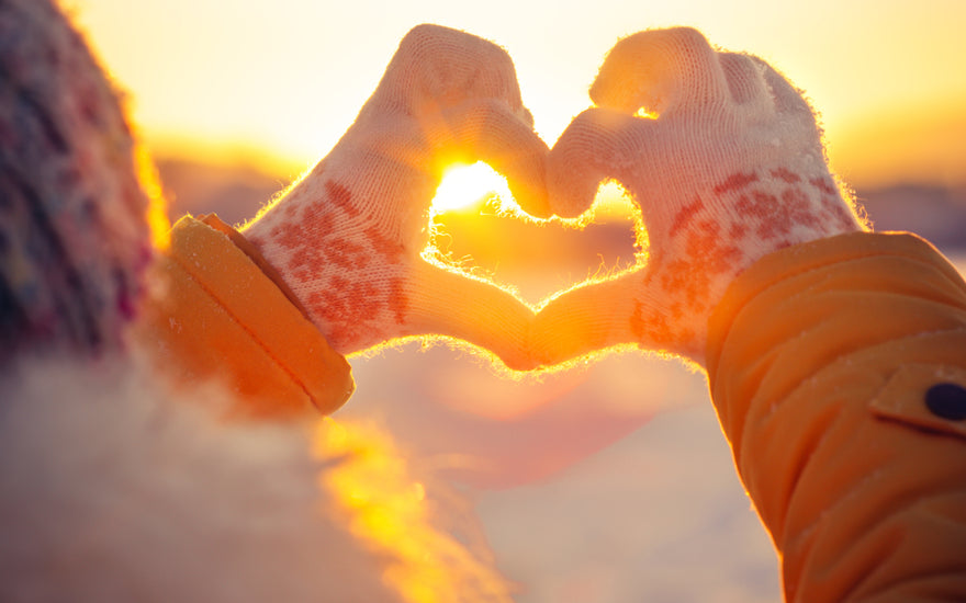 Hands wearing winter gloves are held up with fingers positioned to form a heart in front of the sunset.