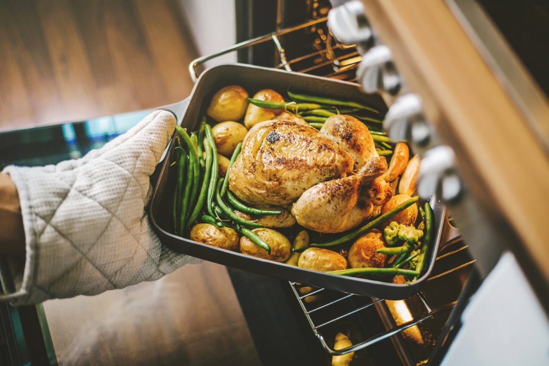 Hands in oven mitts pulling a pan with a turkey, green beans, and potatoes out of the oven. 