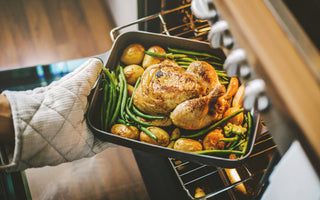 Hands in oven mitts pulling a pan with a turkey, green beans, and potatoes out of the oven. 