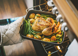 Hands in oven mitts pulling a pan with a turkey, green beans, and potatoes out of the oven. 