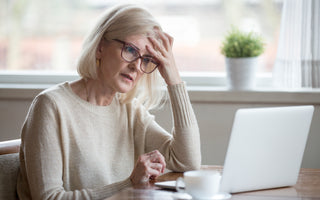 Older woman in a tan sweater sitting in front of an open laptop. She has her head resting on her hand and has a confused look on her face.