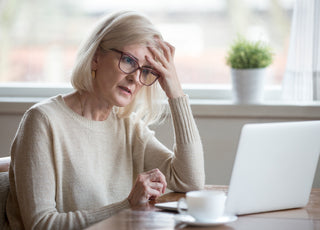 Older woman in a tan sweater sitting in front of an open laptop. She has her head resting on her hand and has a confused look on her face.