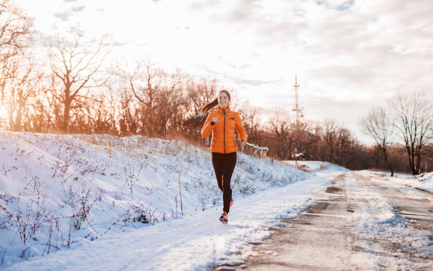 Woman in an orange jacket running along the side of a snowy mountain road. 