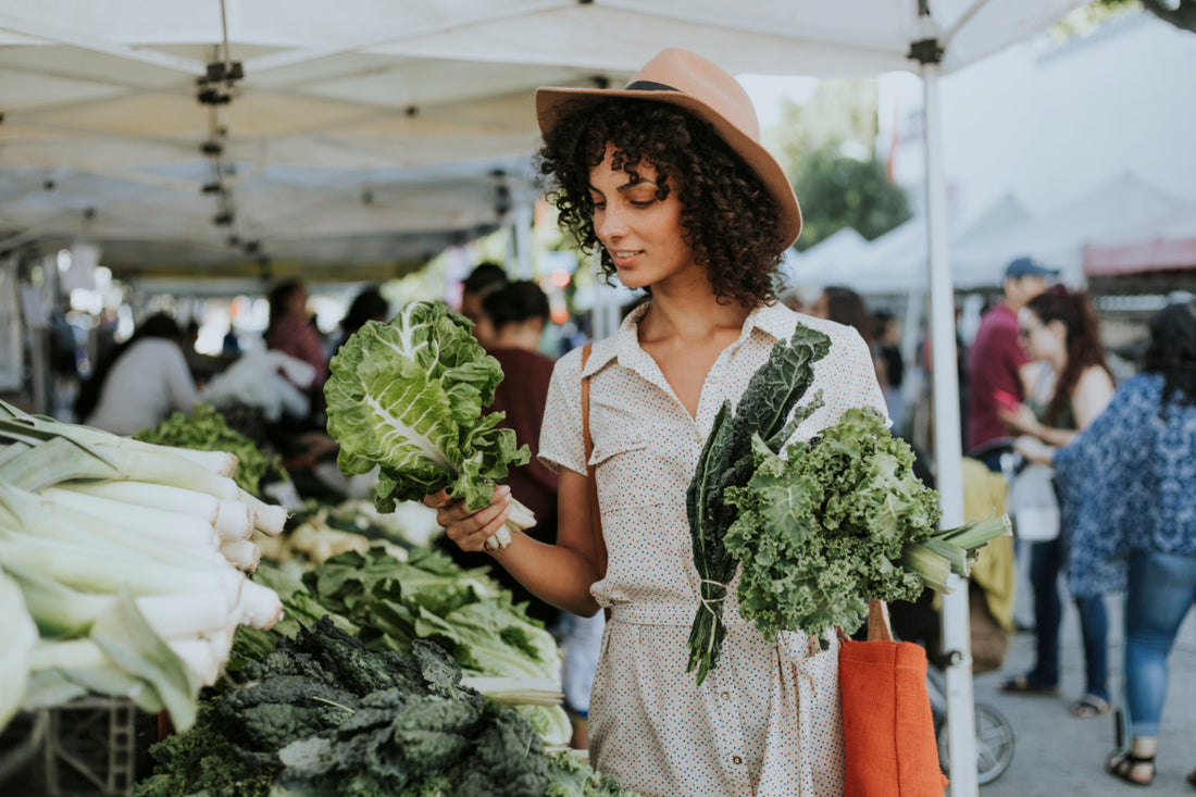 Woman at a farmers market stall examining a selection of lettuce while holding other fresh produce in her left hand.