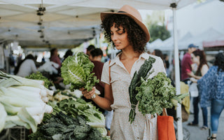 Woman at a farmers market stall examining a selection of lettuce while holding other fresh produce in her left hand.