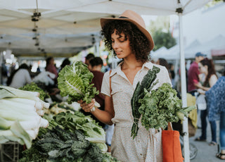 Woman at a farmers market stall examining a selection of lettuce while holding other fresh produce in her left hand.