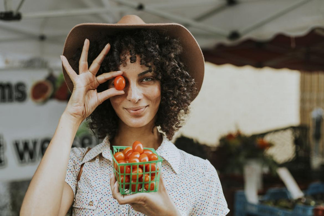 Women in a sunhat holding a small container of cherry tomatoes and holding one of the tomatoes in front of her eye.
