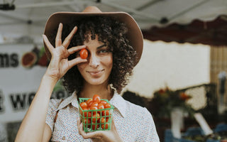 Women in a sunhat holding a small container of cherry tomatoes and holding one of the tomatoes in front of her eye.