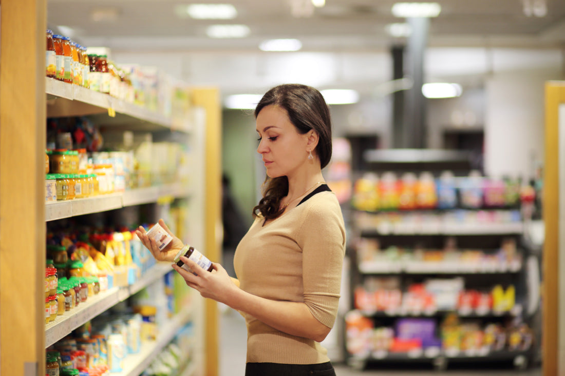 Woman wearing business casual attire standing in a grocery store aisle reading and comparing the labels of two different items.