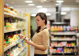 Woman wearing business casual attire standing in a grocery store aisle reading and comparing the labels of two different items.