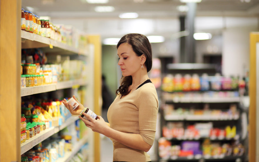 Woman wearing business casual attire standing in a grocery store aisle reading and comparing the labels of two different items.