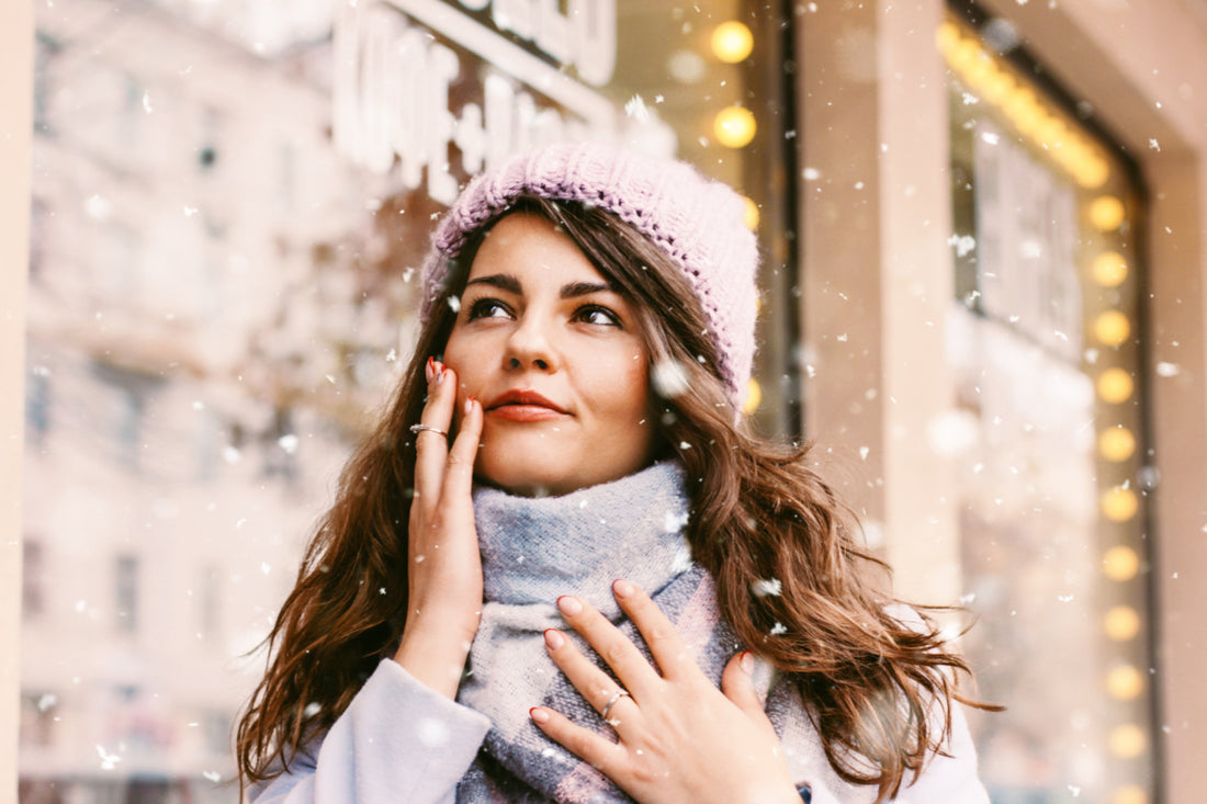 Woman wearing a blue sweater, plaid scarf, and purple beanie standing outside with her fingertips on her feet while it's snowing in the city..