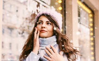 Woman wearing a blue sweater, plaid scarf, and purple beanie standing outside with her fingertips on her feet while it's snowing in the city..