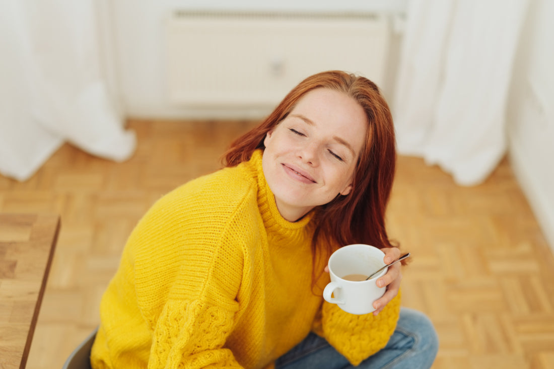 Woman in a yellow sweater sitting cross-legged on a wooden floor holding a cup of tea in her hand with her head tilted up, her eyes closed, and a smile on her face.