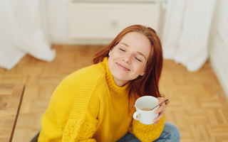 Woman in a yellow sweater sitting cross-legged on a wooden floor holding a cup of tea in her hand with her head tilted up, her eyes closed, and a smile on her face.