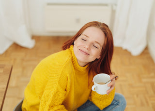 Woman in a yellow sweater sitting cross-legged on a wooden floor holding a cup of tea in her hand with her head tilted up, her eyes closed, and a smile on her face.