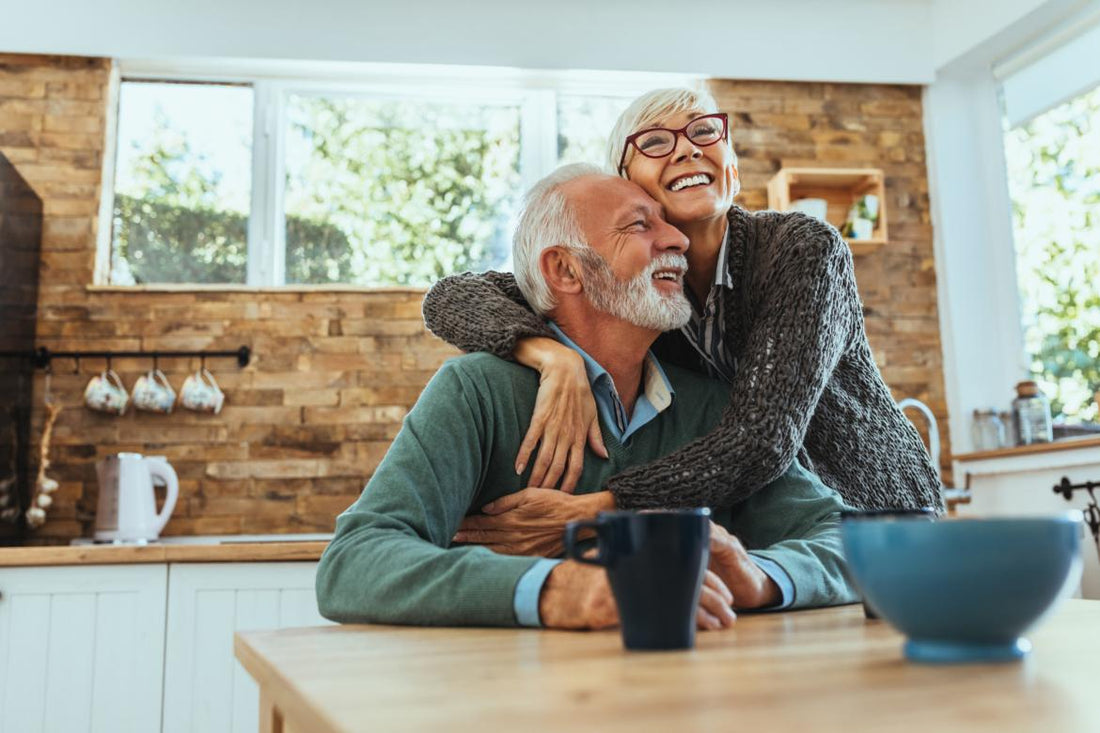 Senior woman hugging a senior man who is sitting at the table in the kitchen.