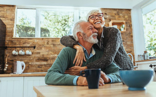 Senior woman hugging a senior man who is sitting at the table in the kitchen.