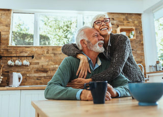 Senior woman hugging a senior man who is sitting at the table in the kitchen.