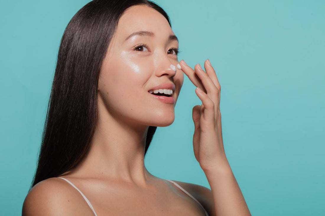 Asian women with dark straight hair applying a white moisturizer to her face in front of a light blue background. 
