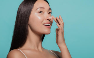 Asian women with dark straight hair applying a white moisturizer to her face in front of a light blue background. 