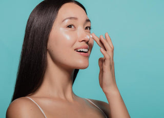 Asian women with dark straight hair applying a white moisturizer to her face in front of a light blue background. 