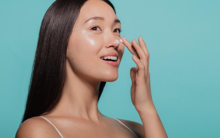 Asian women with dark straight hair applying a white moisturizer to her face in front of a light blue background. 