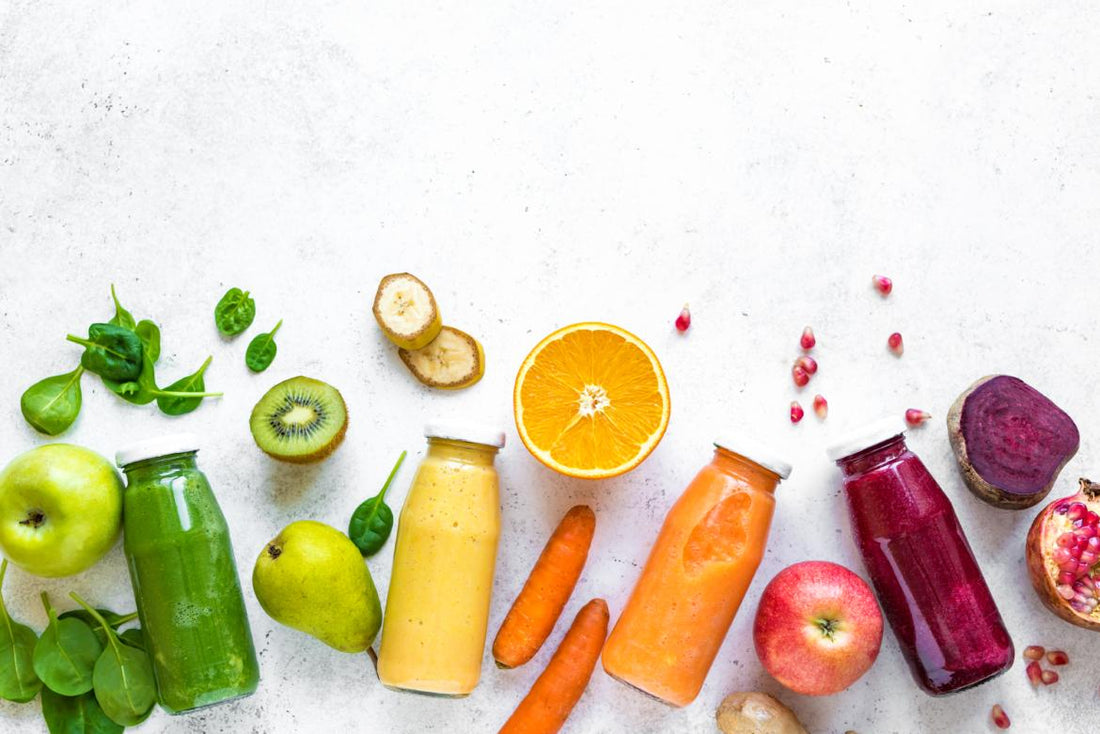 Four bottles of colorful juice laying on a counter surrounded by matching ingredients.