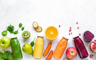 Four bottles of colorful juice laying on a counter surrounded by matching ingredients.