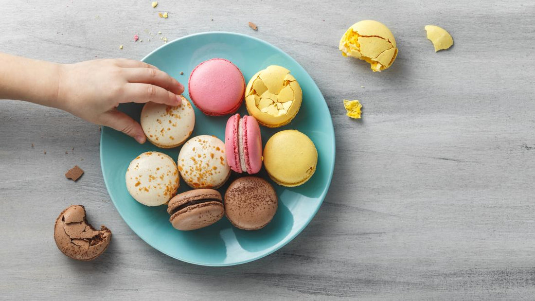 Top down view of color macrons on a blue plate. A child's hand is reaching to grab one of them.