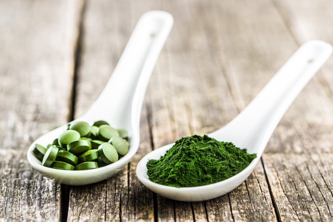 Two white measuring spoons sitting on a wooden table. One is filled with green tablets and the other is filled with green powder.