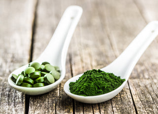 Two white measuring spoons sitting on a wooden table. One is filled with green tablets and the other is filled with green powder.