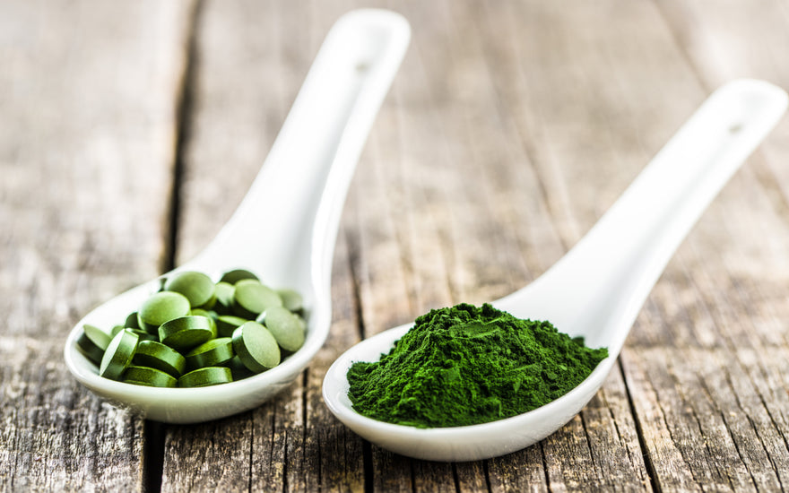 Two white measuring spoons sitting on a wooden table. One is filled with green tablets and the other is filled with green powder.