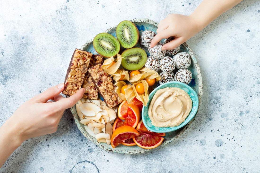 Two hands reaching to grab food off of a stone platter holding a variety of snack items including kiwis, granola bars, and hummus.