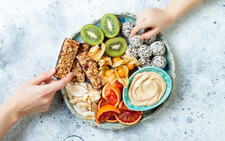 Two hands reaching to grab food off of a stone platter holding a variety of snack items including kiwis, granola bars, and hummus.