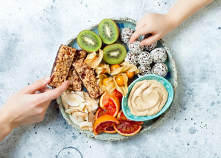 Two hands reaching to grab food off of a stone platter holding a variety of snack items including kiwis, granola bars, and hummus.