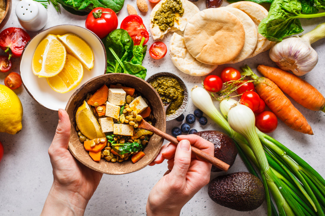 Pair of hands holding a wooden bowl filled with fresh plant-based foods over top of a countertop that is filled with similar food items.