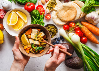 Pair of hands holding a wooden bowl filled with fresh plant-based foods over top of a countertop that is filled with similar food items.