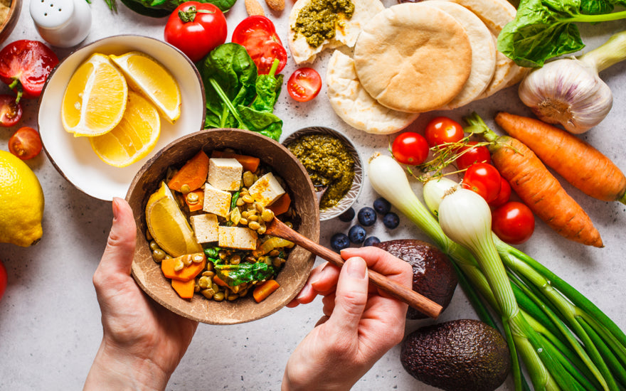 Pair of hands holding a wooden bowl filled with fresh plant-based foods over top of a countertop that is filled with similar food items.